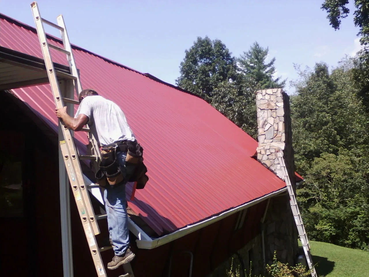 Roofer on a ladder beside a red metal roof and stone chimney — Soffit & Fascia Repair by Roofing Brothers in Gamaliel